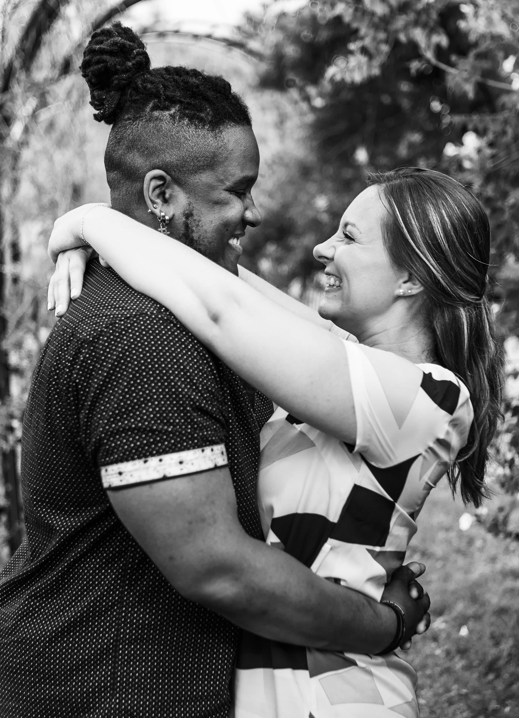 A black and white photo with Jalil Malik on the left and Ana Van Lonkhuyzen on the right, their arms wrapped around each other as they happily look into each other's eyes.